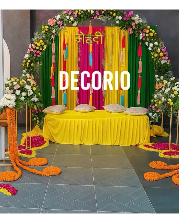 This colorful mehndi seating area features a backdrop of bright pink and green drapes against a floral arch. The low seating with cushions provides a comfortable and traditional space for the bride during the ceremony.