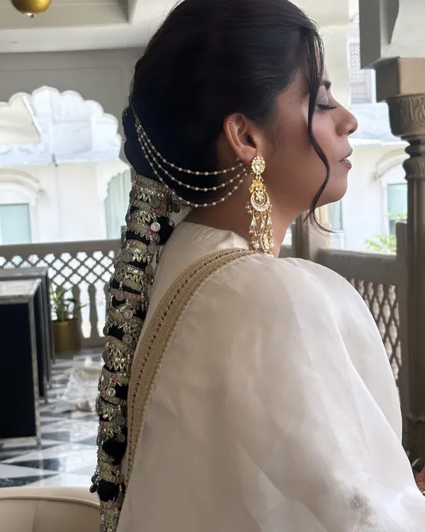 A stunning side profile of an intricate bridal braid. This style features a traditional Punjabi parandi with mirror work and ghungroos, complemented by delicate pearl chains that connect to the earrings. It's a true work of art.