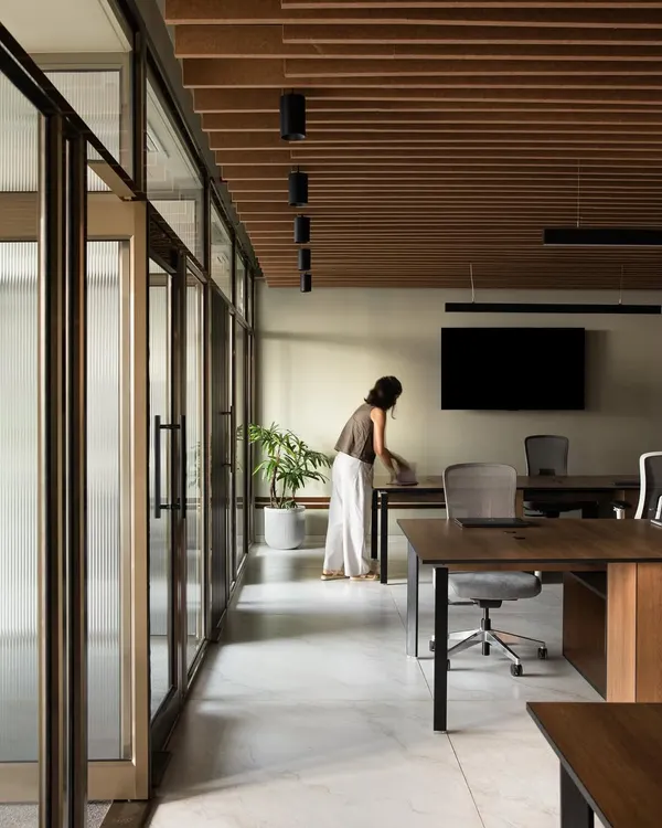 This wide shot of the office shows an open and airy workspace defined by a wooden slatted ceiling and glass partitions. The layout is designed for flexibility, allowing for easy movement and communication between different zones while maintaining a clean, uncluttered aesthetic.