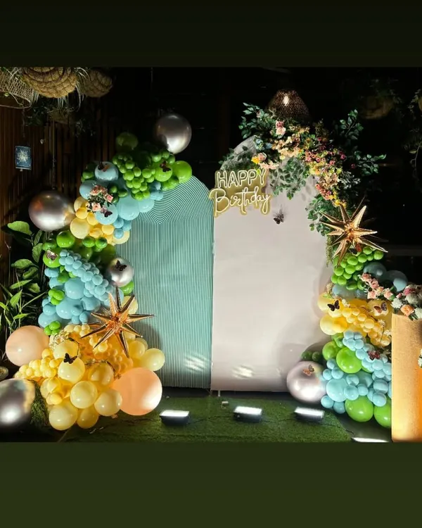 This elegant birthday setup features a unique house-shaped backdrop with floral arrangements. The organic balloon garland in shades of green, yellow, and peach adds a soft and sophisticated touch to the decor.
