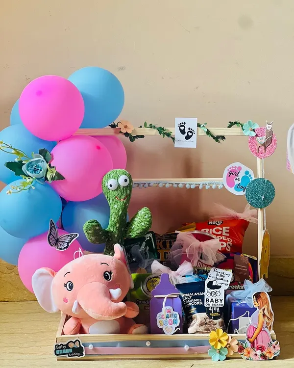 This baby shower hamper is full of fun and surprises. It features a pink elephant and a dancing cactus toy, along with snacks and treats, all set in a decorated wooden cart perfect for a party centerpiece.