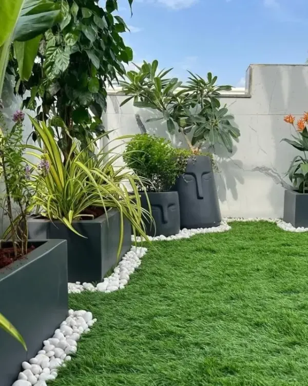 A beautiful terrace garden featuring a mix of artificial turf, modern planters, and white pebble borders. The design incorporates unique face-themed pots and a variety of plants like Birds of Paradise to create a stylish and personalized outdoor space.
