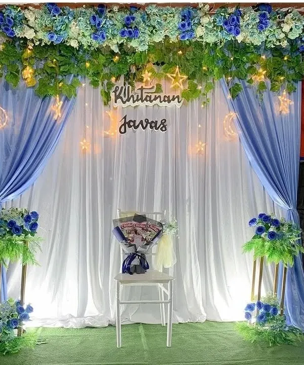 An elegant stage decoration for a 'Khitanan' ceremony, featuring blue and white drapes, floral arrangements, and fairy lights.