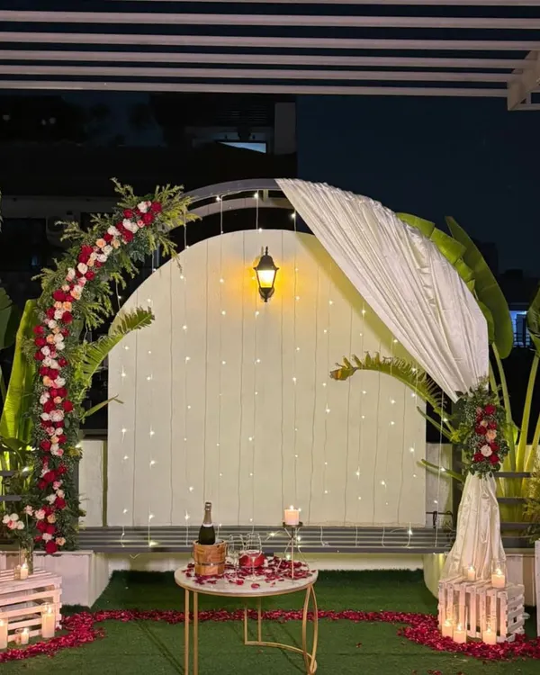 This is the complete romantic terrace setup at night. A floral arch is illuminated by fairy lights, with a candlelit table for two set for a private dinner.