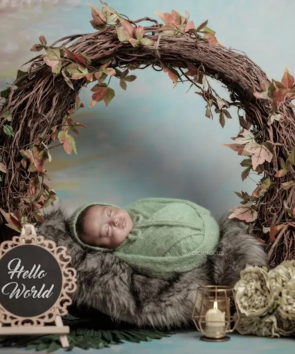 A baby swaddled in green, sleeping peacefully inside a large wreath made of natural vines and leaves. This 'Hello World' setup is a beautiful way to announce your new arrival.