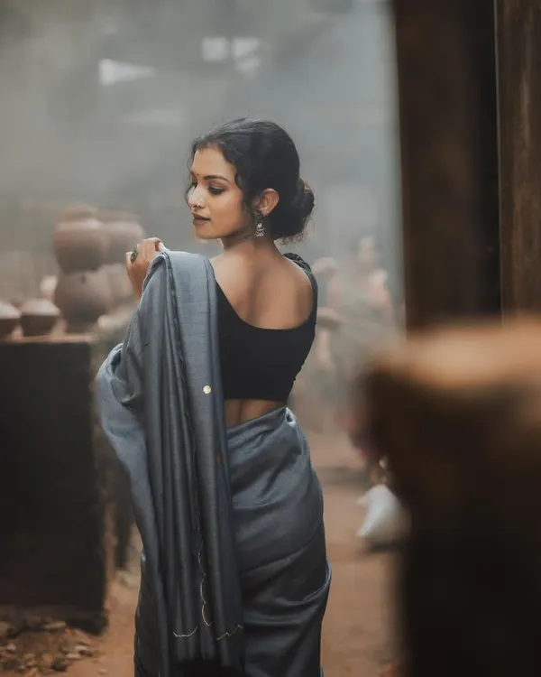 A beautiful back profile shot that creates a sense of mystery and elegance. The model, draped in a simple grey saree, looks over her shoulder in a rustic, hazy setting, highlighting her graceful posture.