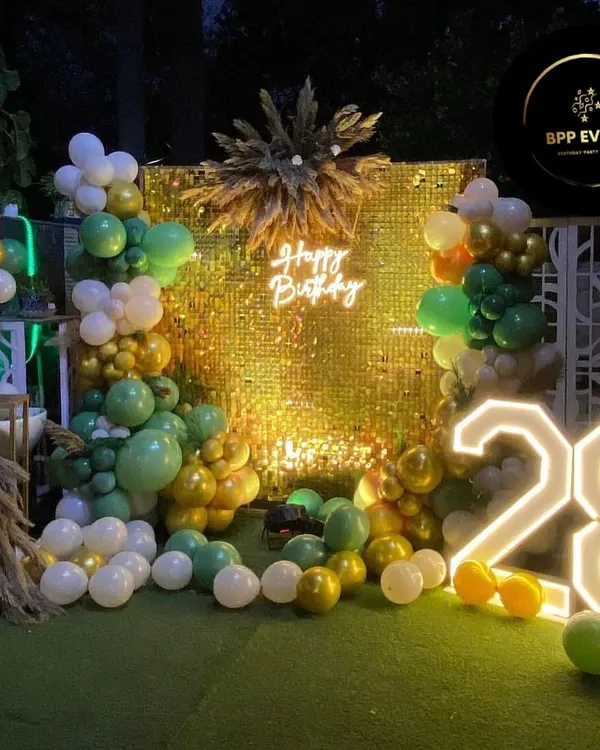 A stunning outdoor birthday celebration setup featuring a golden sequin wall for a glamorous photo backdrop. The look is completed with a 'Happy Birthday' neon sign, large marquee numbers showing the age '28', and an organic balloon arrangement in shades of green, white, and gold.