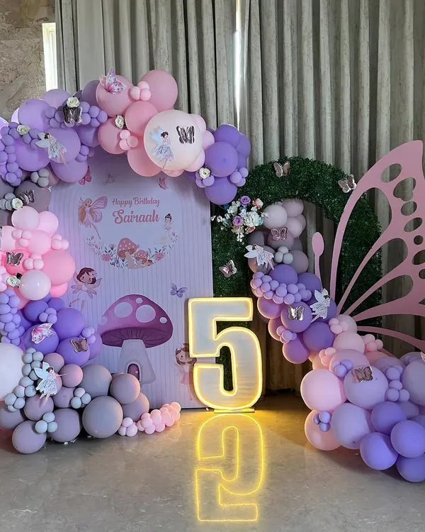 A whimsical fairy and butterfly-themed fifth birthday setup, featuring a large light-up number '5' and a stunning pink butterfly wing prop. The scene is completed with a custom backdrop of an enchanted forest and a pastel balloon arch in pink, purple, and grey.