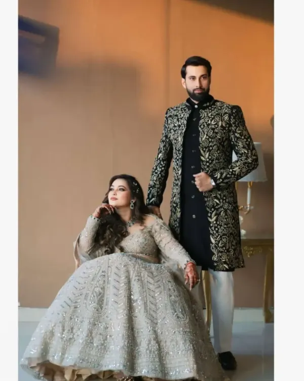 A stunning couple's portrait. The groom wears our black velvet Indo-Western sherwani with intricate all-over golden embroidery, paired with classic white trousers for a look of pure opulence.