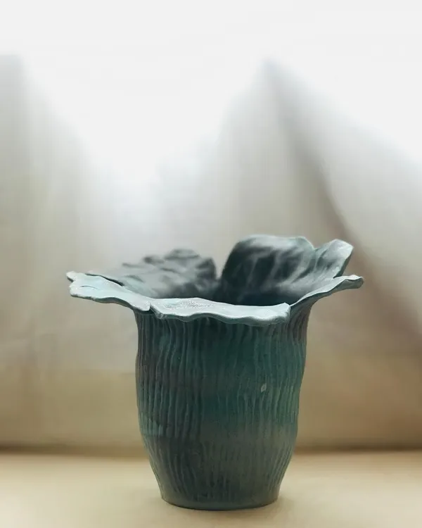 The Wild Petal Vase, a piece of functional sculpture. The hand-shaped rim mimics the organic form of a flower, and the textured, dark green glaze gives it a deep, earthy feel.
