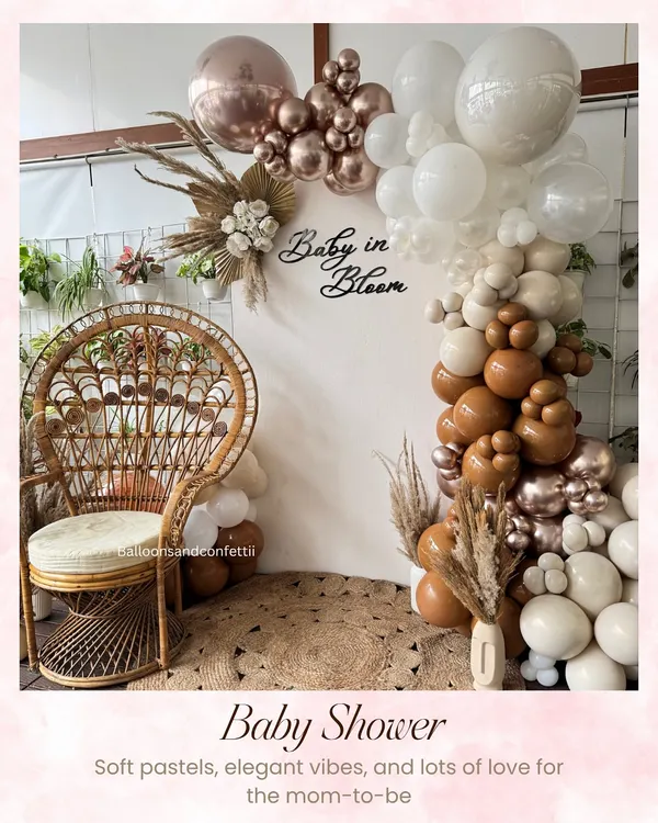 A soft and elegant 'Baby in Bloom' baby shower setup. The neutral-toned balloon arch, pampas grass, and wicker peacock chair create a warm and stylish vibe for the mom-to-be.
