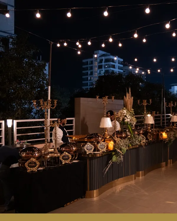 An elegant buffet line under a canopy of string lights. The combination of dark, modern counters, gold chafing dishes, and classic table lamps creates a sophisticated and inviting atmosphere for an evening event.