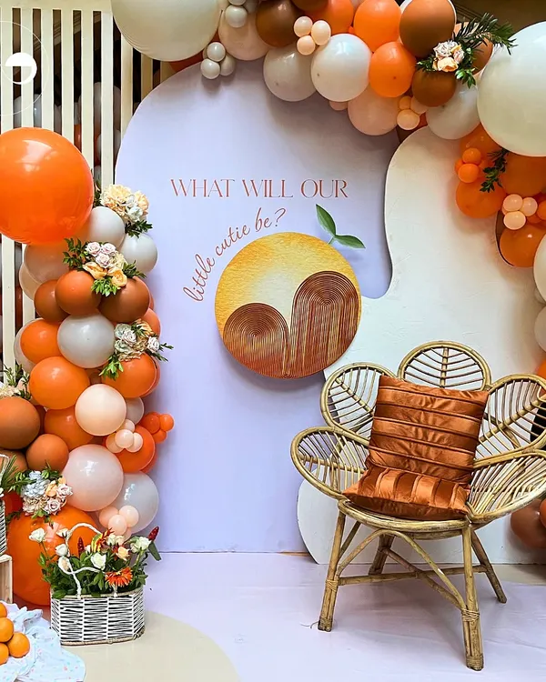 This "Little Cutie" gender reveal setup combines a playful theme with chic design. A custom backdrop, a stylish rattan chair, and a vibrant orange and white balloon arch create a stunning focal point for photos and fun.