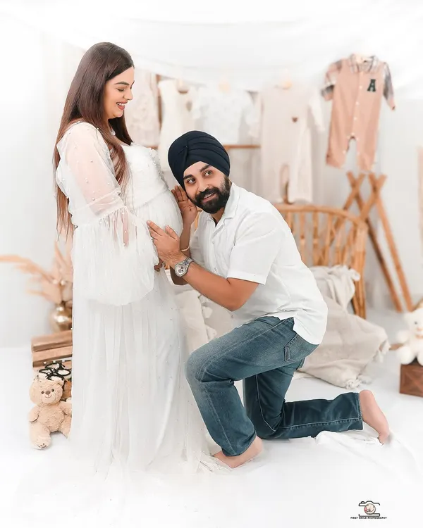 A joyful dad to be listens to the baby bump during a studio maternity shoot. The background is styled with neutral baby clothes, creating a warm and loving scene.