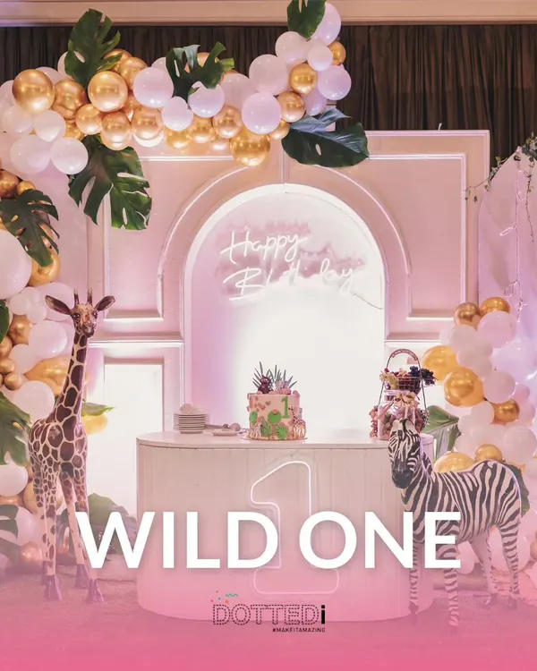 A "Wild One" jungle-themed first birthday party setup. The decor features a beautiful balloon arch in white and gold, life-sized giraffe and zebra figures, and a custom cake table.
