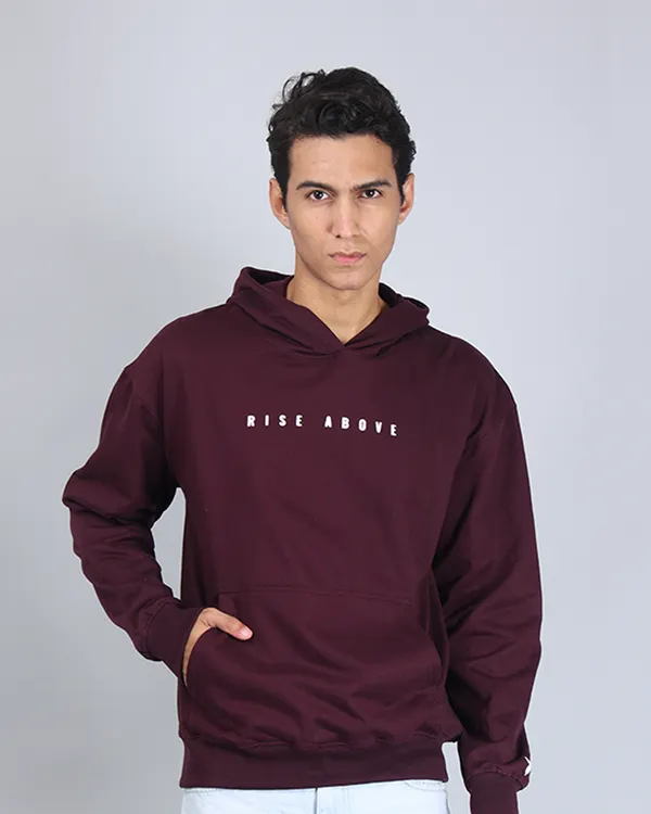 A confident, front-facing shot of a model in a maroon "Rise Above" hoodie. The clean grey background ensures the focus remains entirely on the product.