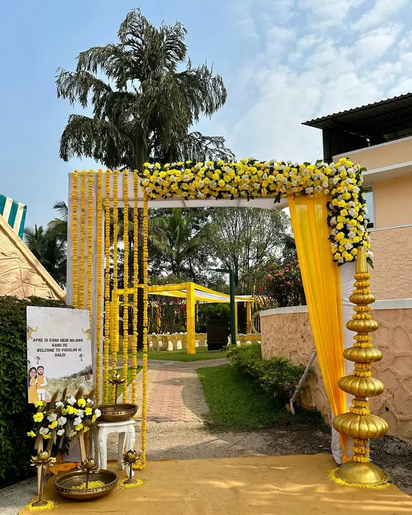 A bright and welcoming entrance for a Haldi ceremony, featuring a simple arch decorated with yellow marigold garlands and a custom welcome sign. The golden temple-style pillar adds a touch of traditional elegance to the outdoor venue.