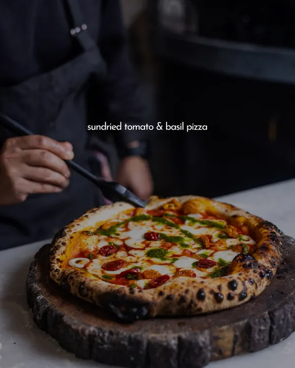 This is what it's all about. A beautiful Sundried Tomato & Basil pizza, fresh from the wood-fired oven. You can see the perfectly charred crust, the melted mozzarella, and the vibrant pesto, all crafted by our pizzaiolo right before your eyes.