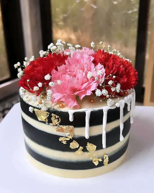 A striking black and white striped cake with a white chocolate drip, gold leaf, and a beautiful arrangement of fresh carnations. This design is modern, bold, and elegant.