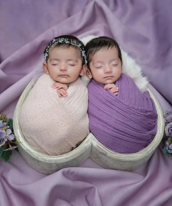 A beautiful portrait of newborn twins swaddled in pink and purple, sleeping peacefully side-by-side in a heart-shaped basket against a lavender backdrop.