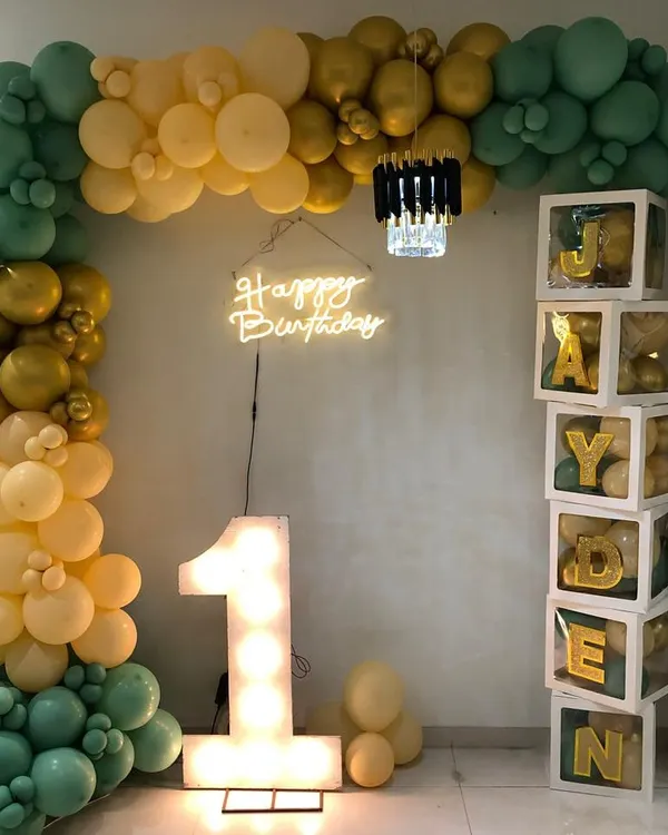 A stylish and modern first birthday for Jayden. This setup features a beautiful balloon arch in sage green, gold, and cream, a light-up number '1', a neon sign, and custom blocks spelling out his name.