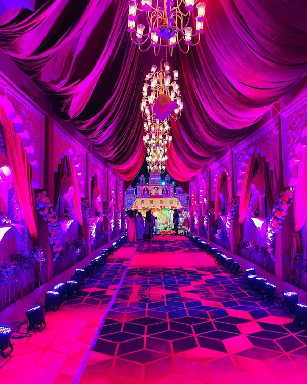 A royal welcome for a grand wedding. This long, majestic entryway features dramatic velvet draping, geometric-patterned carpeting, and stunning chandeliers, all illuminated with vibrant magenta lighting.
