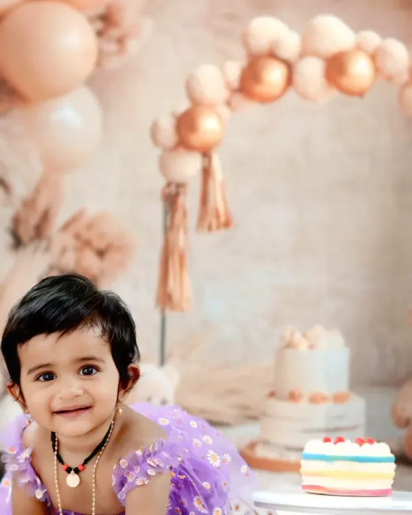 A beautiful baby girl with a radiant smile, captured during her cake smash session with a pastel balloon arch in the background.