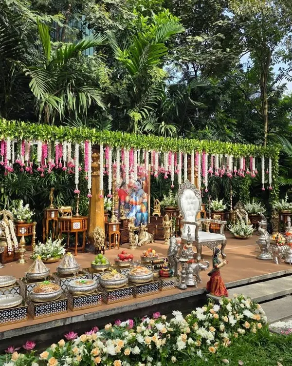A beautiful outdoor stage set against a lush green backdrop of trees. The decor features a canopy of leaves with hanging pink and white garlands, and a Ganesha idol at the center.