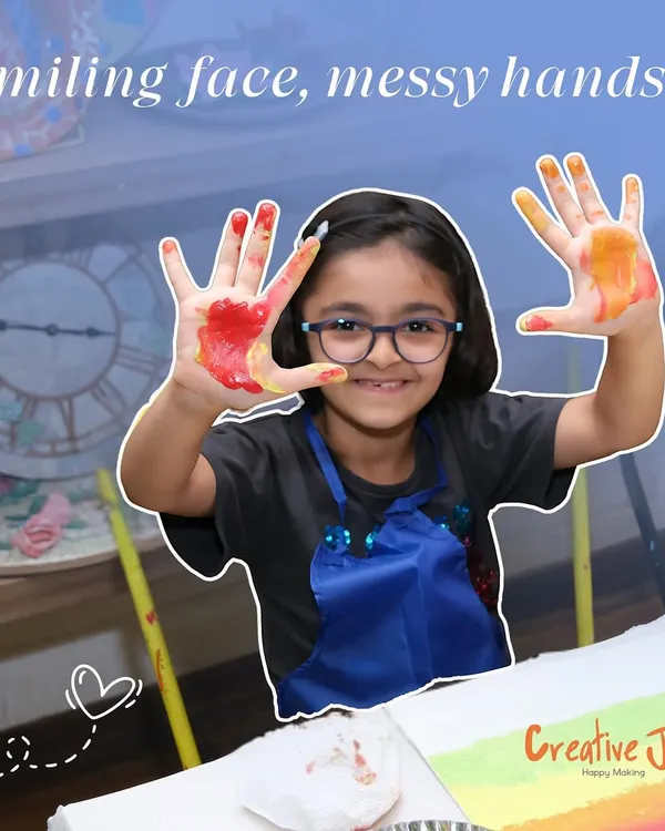 A young girl with glasses smiles, showing off her paint-covered hands. This is the sign of a fun and successful art session.