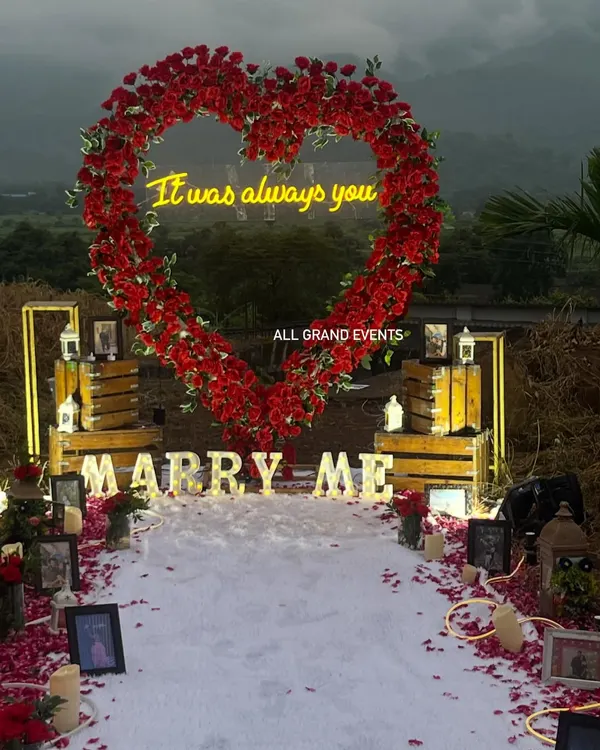 For a truly unforgettable moment, we designed this grand outdoor proposal setup. The centerpiece is a stunning heart-shaped arch made of hundreds of red roses, with a custom neon sign saying 'It was always you'. The aisle is lined with a white carpet, scattered rose petals, and framed photos, creating a personalized journey to the big question.