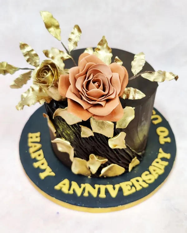 This anniversary cake is a statement of opulent romance, featuring a rich, dark chocolate exterior with a textured finish. A handcrafted sugar rose in a muted blush tone is complemented by gilded leaves and shimmering gold leaf fragments, creating a luxurious and timeless design.