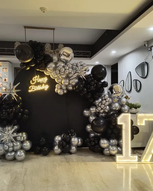 A sleek and modern setup for a 17th birthday, featuring a bold black backdrop framed by a silver and black organic balloon arch. The design is completed with a custom 'Happy Birthday' neon sign and large, light-up marquee numbers, creating a sophisticated party atmosphere.