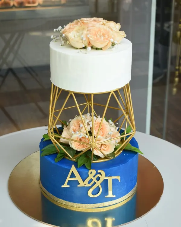 This two-tier cake features a bold royal blue base with custom gold monogramming, separated by a geometric gold cage holding fresh peach roses. The top tier is a clean, elegant white, creating a modern and striking look for a truly special day.