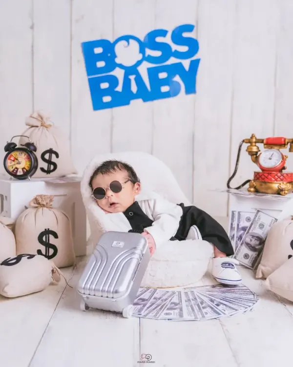 A series from a 'Boss Baby' themed newborn shoot. The setup includes a tiny armchair, a miniature briefcase, and prop money, creating a playful and humorous scene for a baby boy's first portraits.