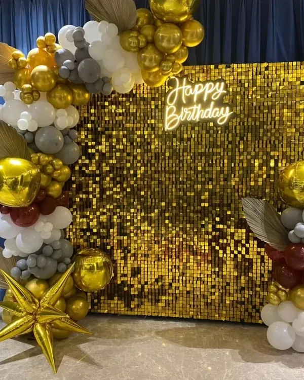 A beautiful and modern birthday backdrop featuring a gold shimmer wall, a custom neon sign, and an organic balloon garland. This setup is perfect for creating a dedicated photo area at any office party.