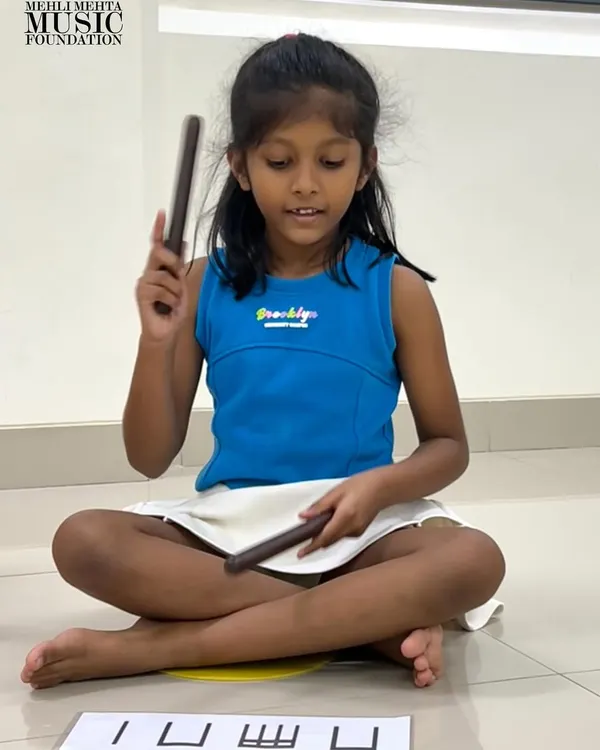 A student uses claves to tap out a rhythm shown on a flashcard. This exercise directly connects visual notation with auditory and kinesthetic learning, reinforcing the concept of rhythm in a multi-sensory way.