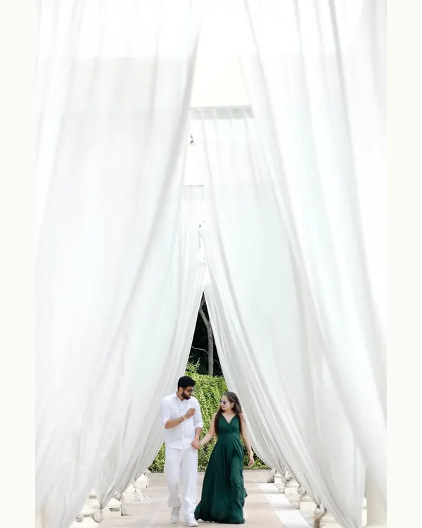 A stunning pre-wedding shot of a couple walking through a corridor of flowing white curtains. This setup creates a dreamy and elegant atmosphere, perfect for a romantic and visually striking photograph.
