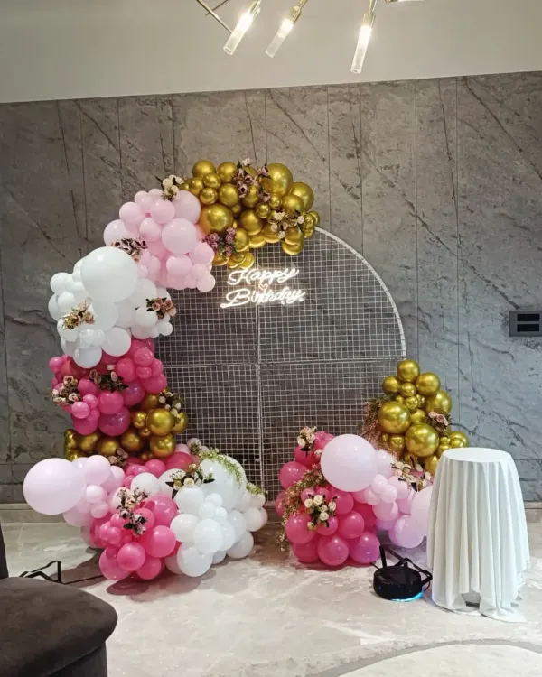 A beautiful circular mesh backdrop decorated with a pink, white, and gold balloon garland. This chic setup includes a 'Happy Birthday' neon sign and is perfect for photos.