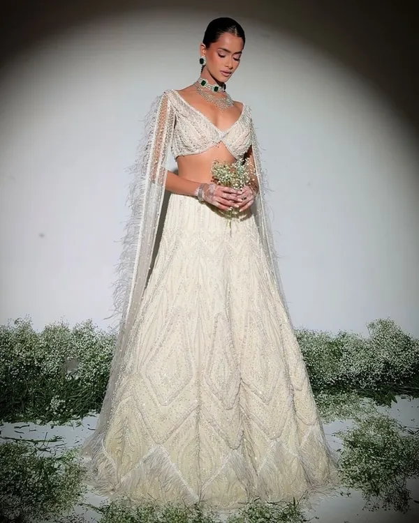 Embodying timeless elegance, this pristine ivory lehenga is embellished with delicate pearls and intricate craftsmanship. The look is completed with feather-trimmed cape sleeves for a touch of modern grace.