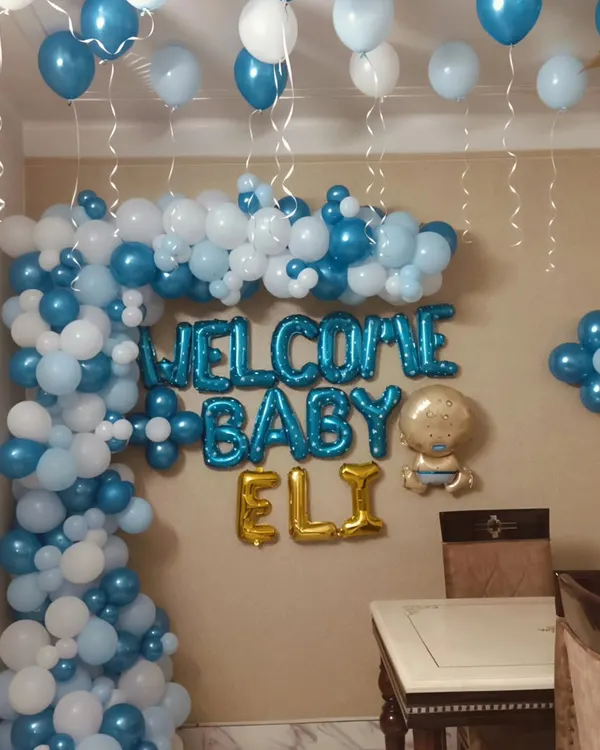 Welcome home, Baby Eli! A classic and joyful home decoration featuring a blue and white balloon semi-arch, 'WELCOME BABY ELI' foil letters, and helium balloons on the ceiling to celebrate the new arrival.
