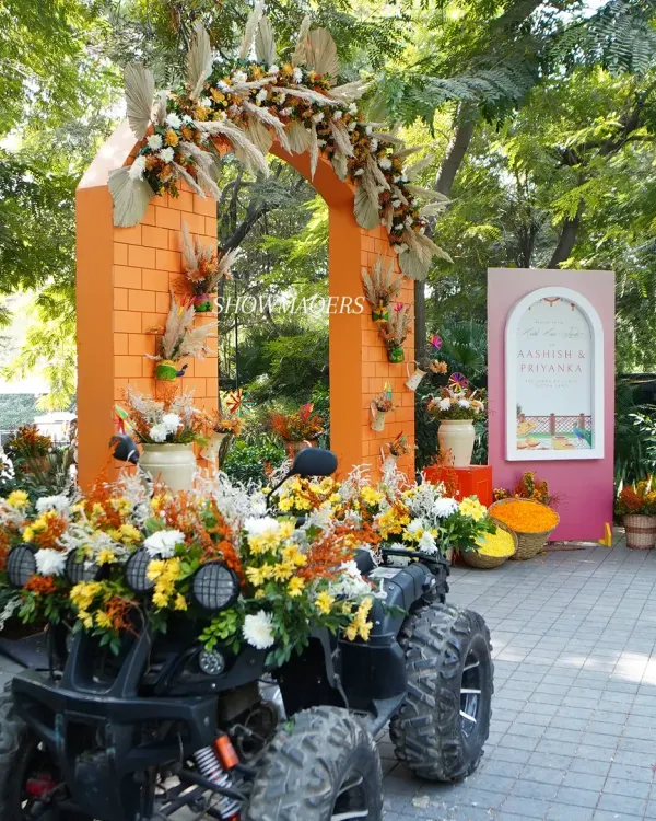 A grand entrance for an orange-themed Haldi, featuring a flower-adorned ATV for the groom's entry. The orange brick-textured arch is decorated with pampas grass and marigolds.