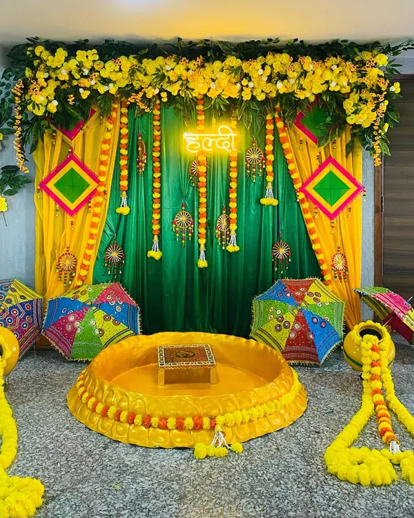 A complete Haldi ceremony setup with a traditional 'urli' for the rituals. The backdrop features yellow and green drapes, marigold garlands, and Rajasthani umbrellas.