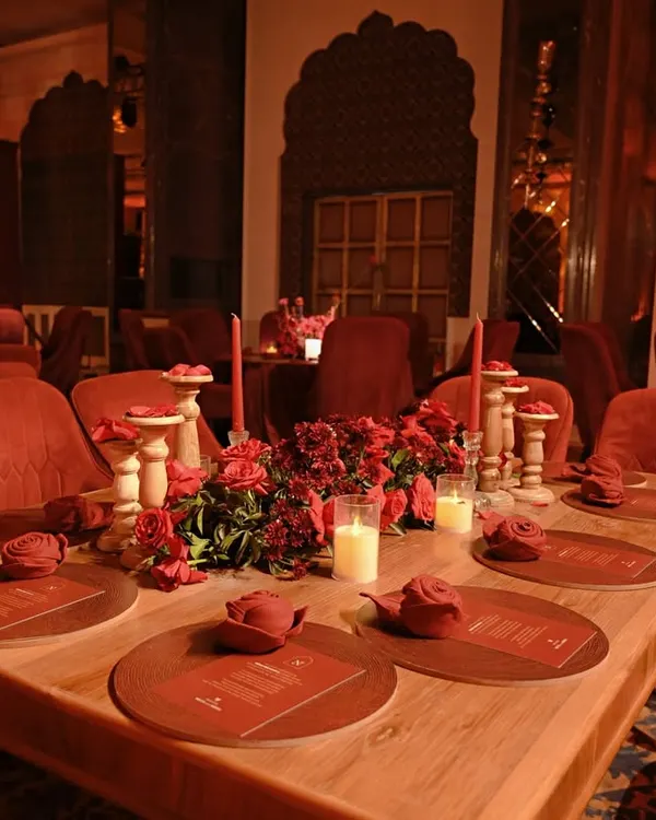 A long wooden table set for an intimate dinner, surrounded by plush red velvet chairs. The centerpiece of red roses and candles creates a warm and romantic focal point.