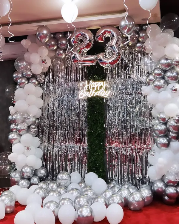 For a stylish 23rd birthday, I created this modern backdrop using a silver and white balloon garland. The design is complemented by a silver shimmer curtain and a custom 'Happy Birthday' neon sign, creating a perfect photo corner for the celebration.