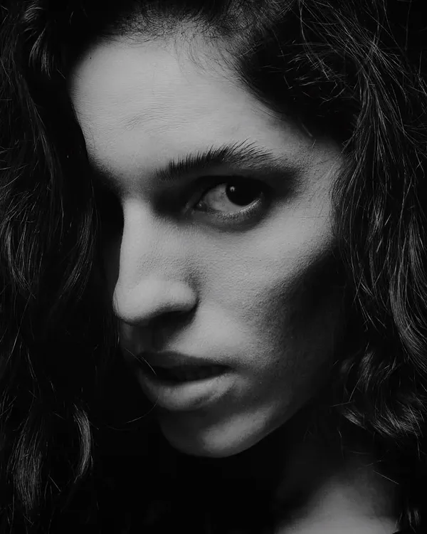An intense black and white close-up. The single light source creates a dramatic shadow across the face, highlighting the eye and creating a sense of mystery and power.