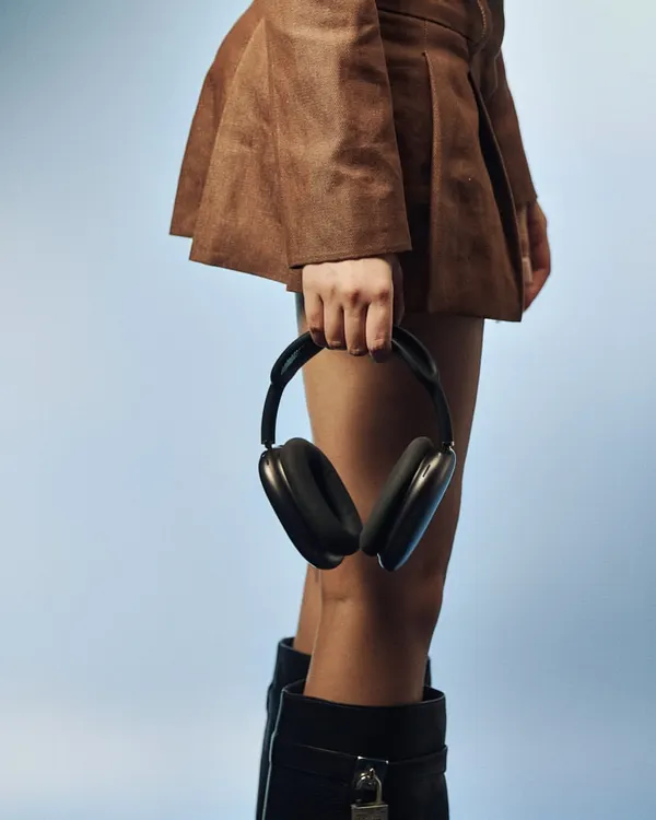 A clean, modern product-in-context shot. The focus is on the sleek design of the black headphones, held against a simple brown skirt and blue background. This style is perfect for e-commerce and social media campaigns for modern accessories.