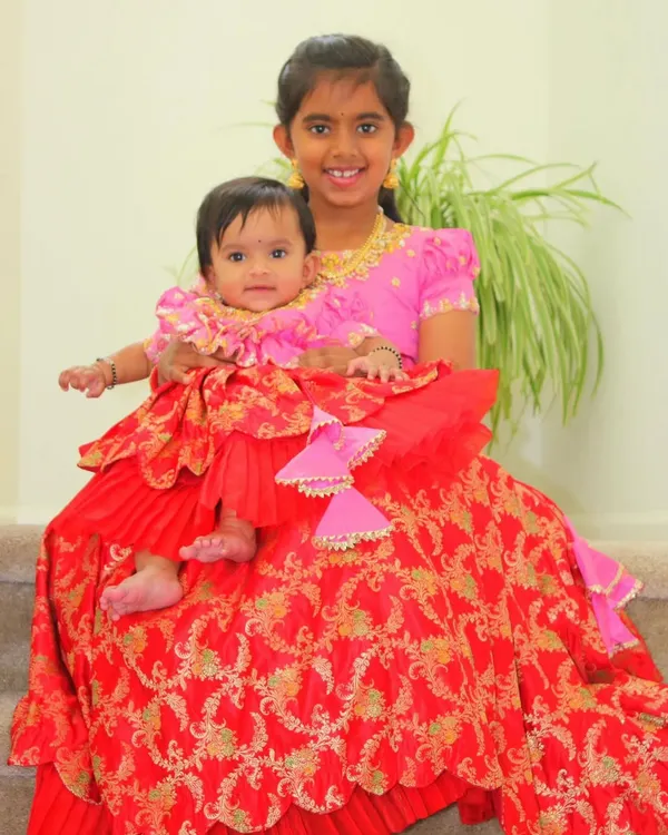Twinning sisters, one big and one small, in our custom-made outfits. The older sister wears a pink blouse, while the baby wears a matching layered pattu pavadai in a vibrant red Banarasi silk.
