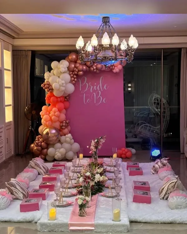 A stunning "Bride to Be" setup for an intimate bridal shower. This luxurious floor seating arrangement features a vibrant pink backdrop, a beautiful organic balloon arch, elegant floral runners, and personalized gift boxes for each guest.