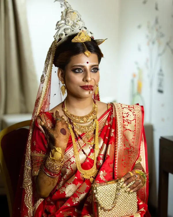 A stunning portrait of a Bengali bride in her traditional red Benarasi saree, adorned with a 'mukut' and intricate gold jewelry.