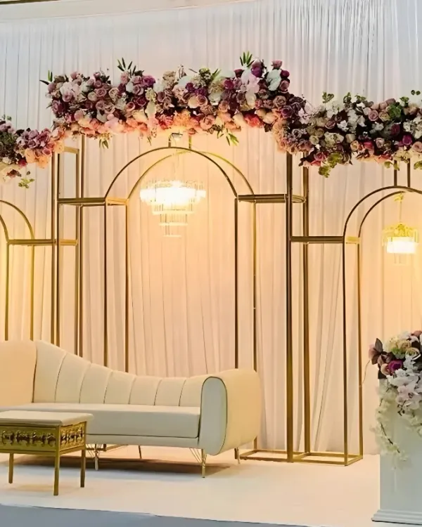 Here is a clear view of a modern and elegant stage I designed. The setup features geometric gold frames, a chic white lounge sofa, and a beautiful arch of purple and pink flowers. A delicate chandelier hangs in the center, adding a touch of classic luxury to the contemporary design.